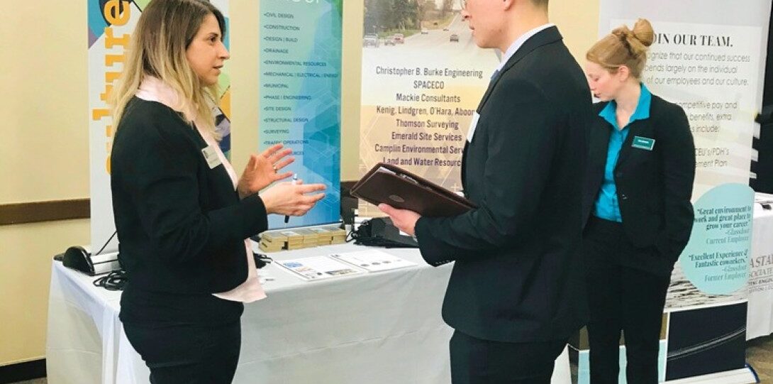 ITE Student Day 2019 Career Fair - A woman on the left in a black suit with a name tag is speaking to a man in a black suit on the right who is holding a brown folder. Another woman in a blue shirt with a name tag is standing in the background.