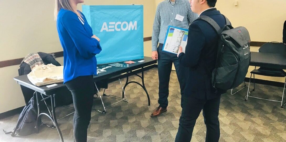 ITE Student Day 2019 Career Fair - Three people stand and converse near an AECOM display table in a well-lit room with beige walls and patterned carpet.