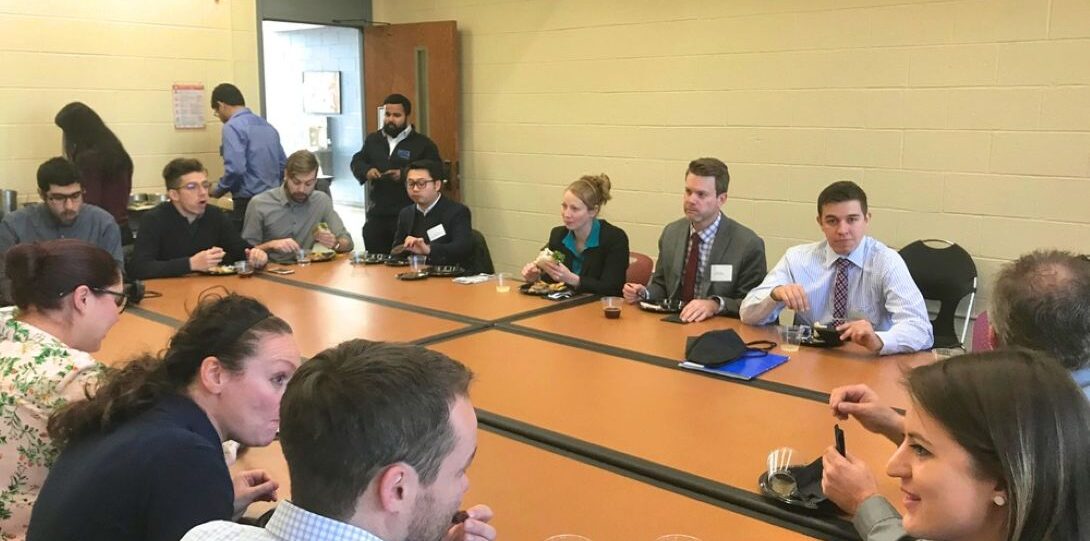 ITE Student Day 2019 - People seated around a conference table eating and conversing in a meeting room with beige walls and a clock on the wall.