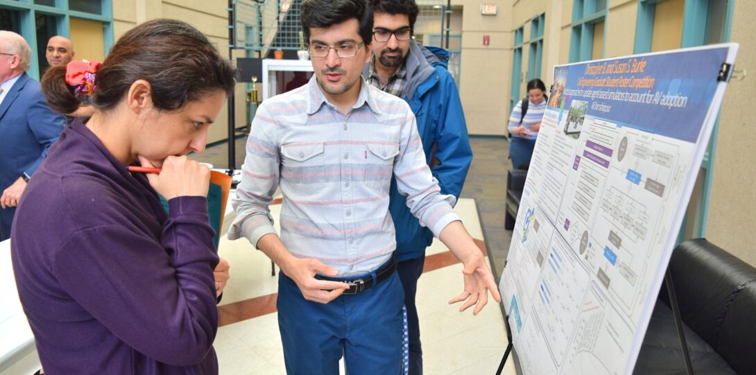 ITE members participating in the CME Poster competition - A poster session where a man in a gray and white striped shirt is pointing at a research poster on an easel and explaining it to a woman in a purple sweatshirt.